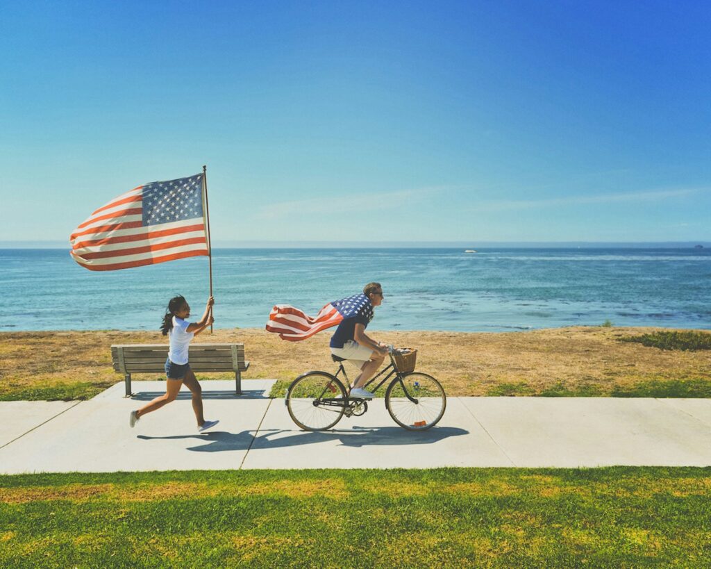 A young man and woman run and ride a bike down the San Diego waterfront, waving American flags.